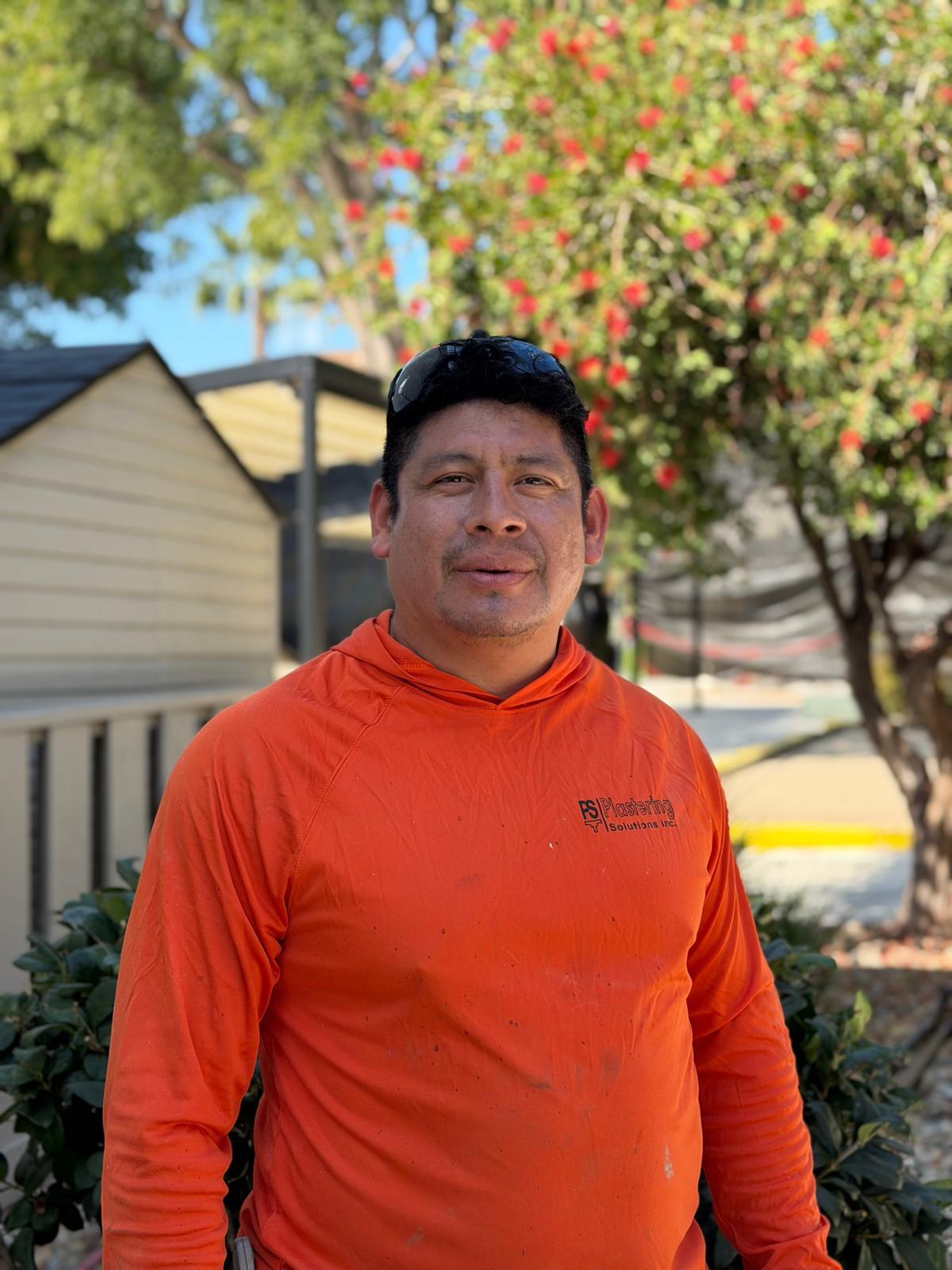 Rivelino Recinos in an orange Plastering Solutions Inc hooded shirt and sunglasses on his cap, standing in front of a flowering tree on a jobsite