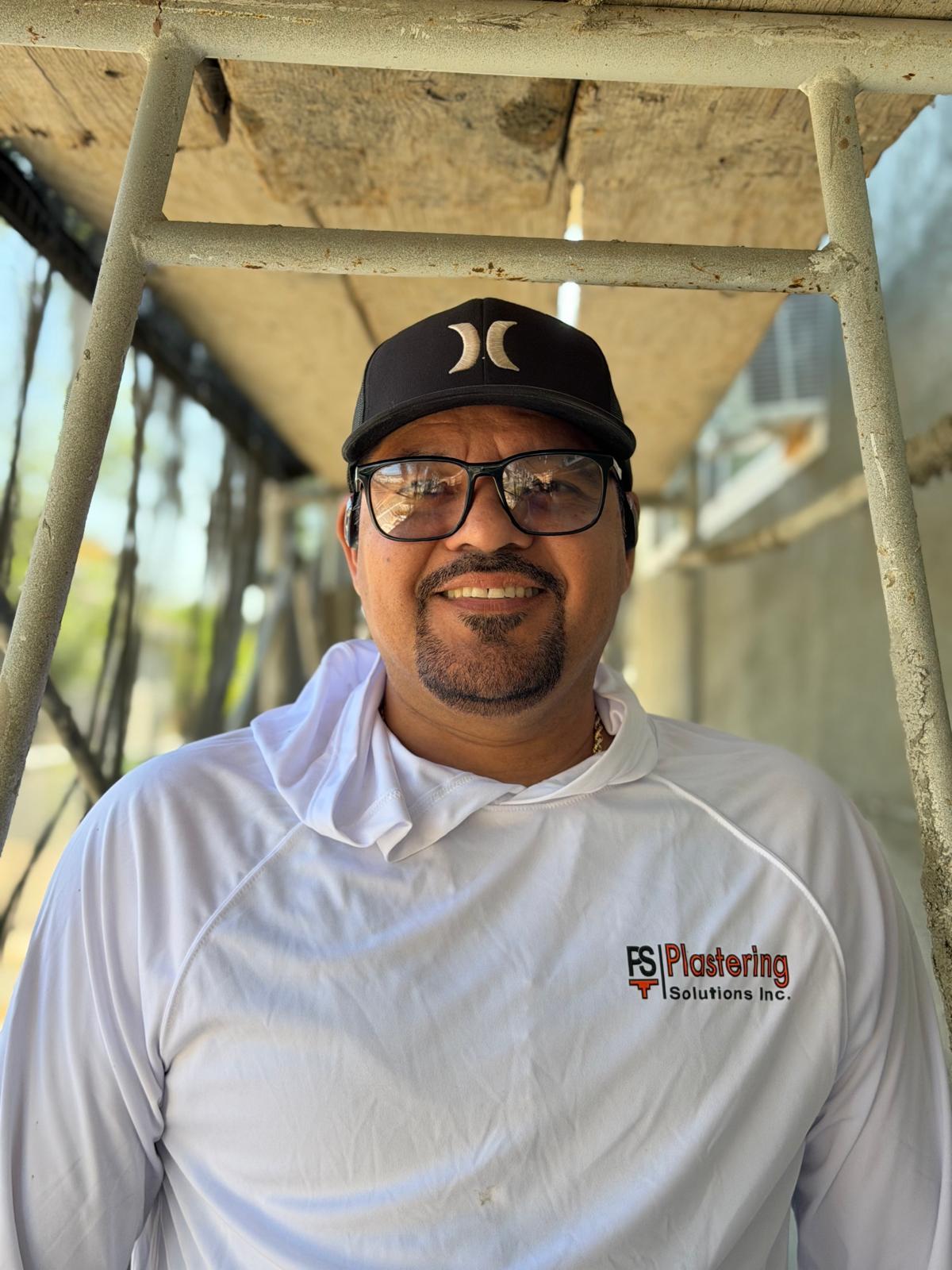 Luis Zamudio in a white Plastering Solutions Inc hooded shirt, glasses and a black cap, standing beneath scaffolding