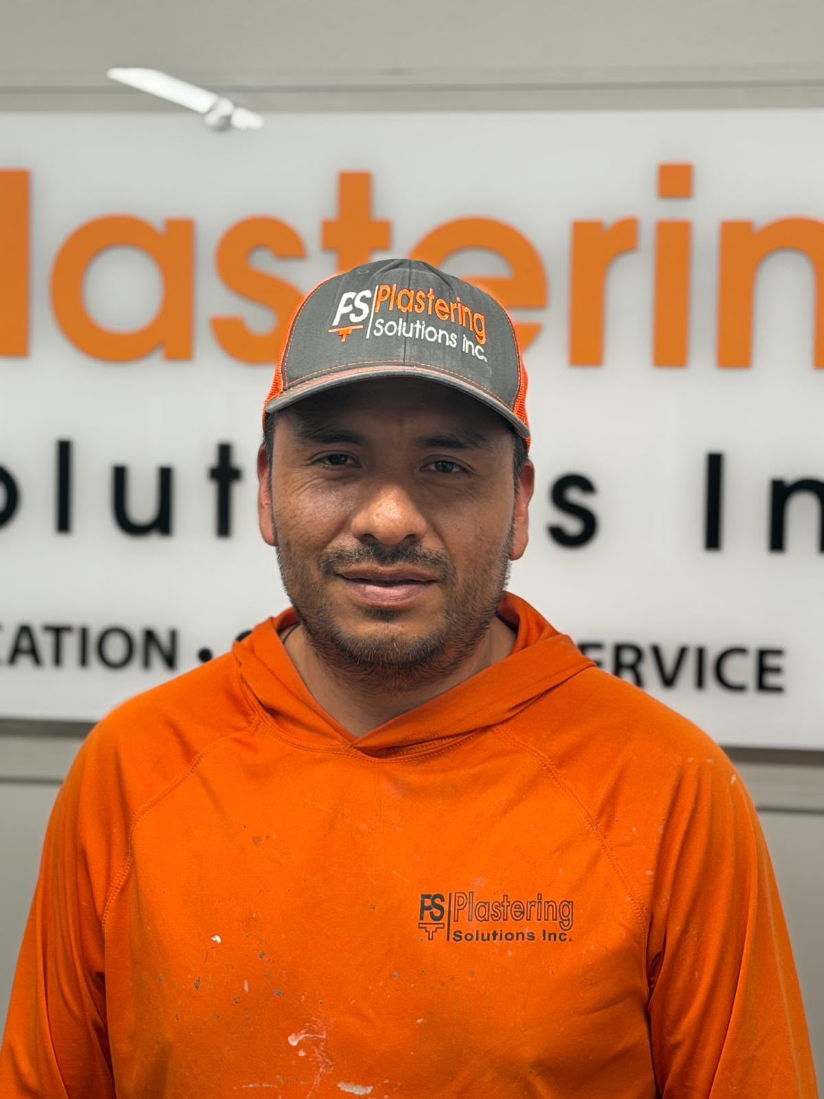 Tomas Martinez in an orange paint speckled Plastering Solutions Inc hooded shirt and cap, in front of the Plastering Solutions Inc office sign