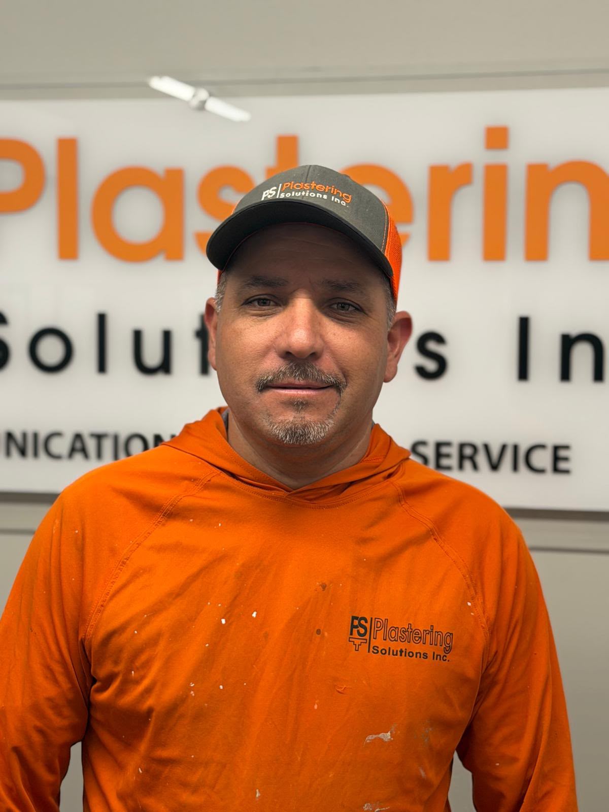 Jose Lopez in an orange paint speckled Plastering Solutions Inc hooded shirt and cap, in front of the Plastering Solutions Inc office sign
