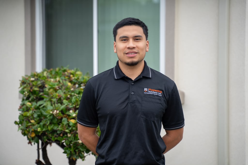 Anthony Rivera in a black Plastering Solutions Inc polo shirt, hands behind his back, beside a trimmed shrub in front of a stucco building