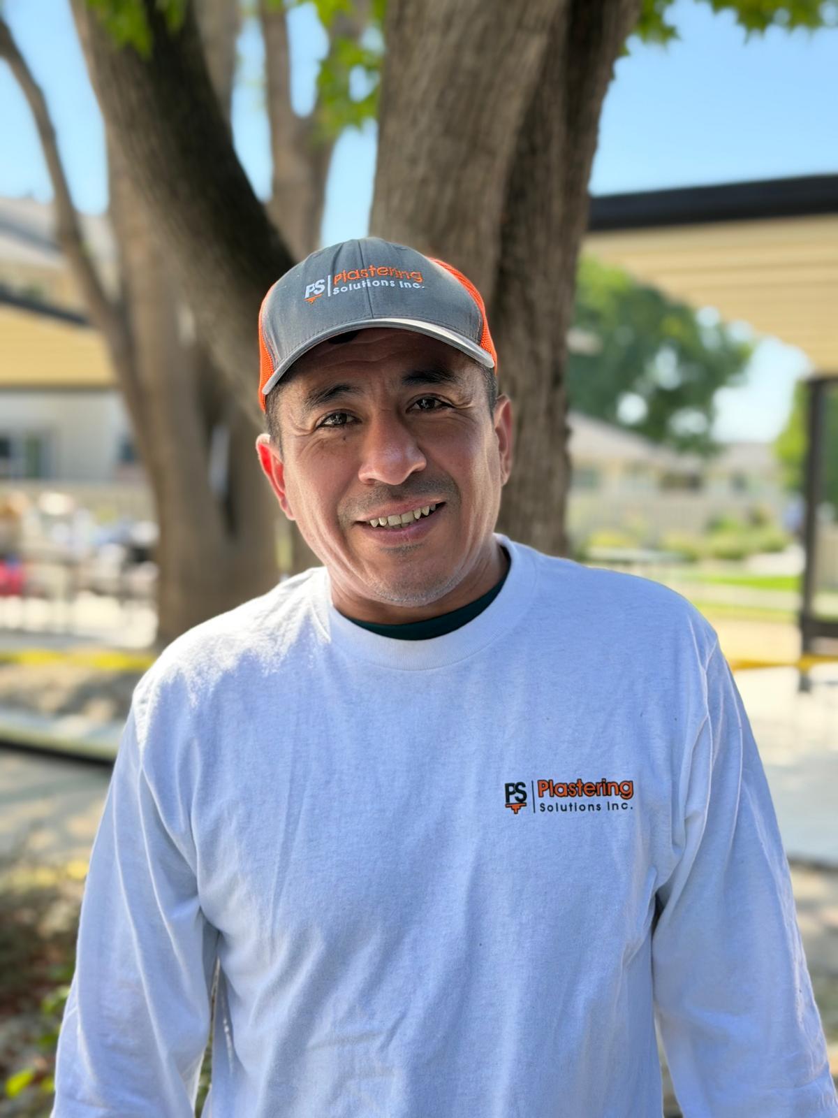 Manuel Lopez in a white long sleeve Plastering Solutions Inc shirt and gray cap, smiling under a shade structure on a jobsite