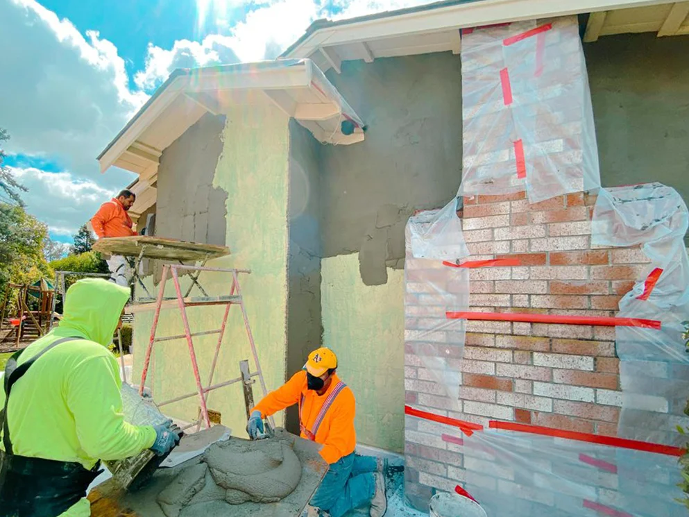 Crew applying new stucco coats while the brick chimney stays protected