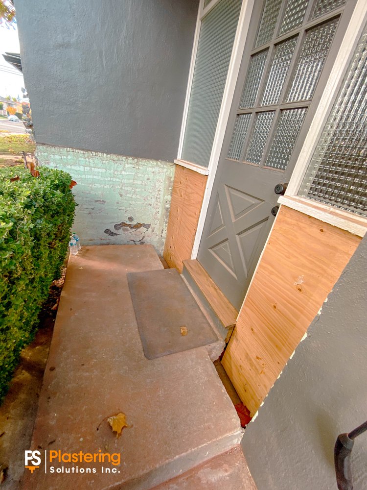 Entry with plywood sheathing beside the door and damaged stucco at the base of the wall