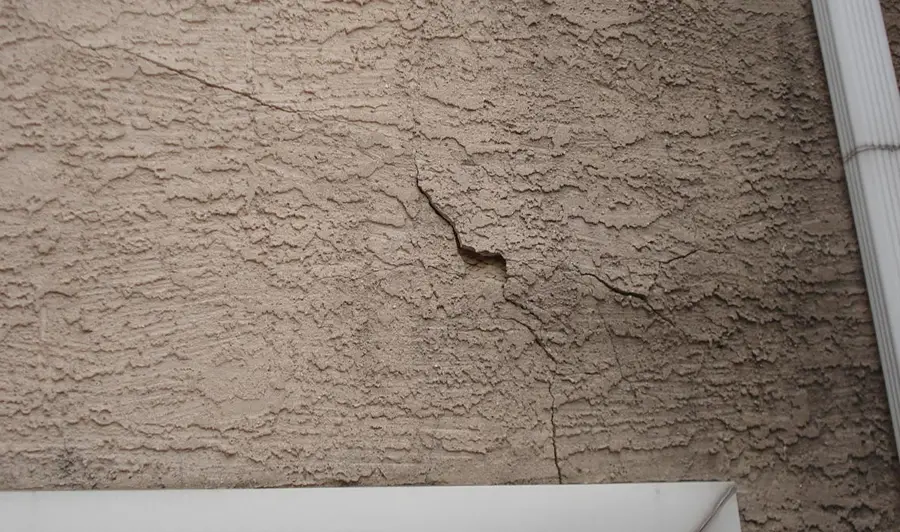 Tan textured stucco wall with a widening crack and dark discoloration beside a white downspout