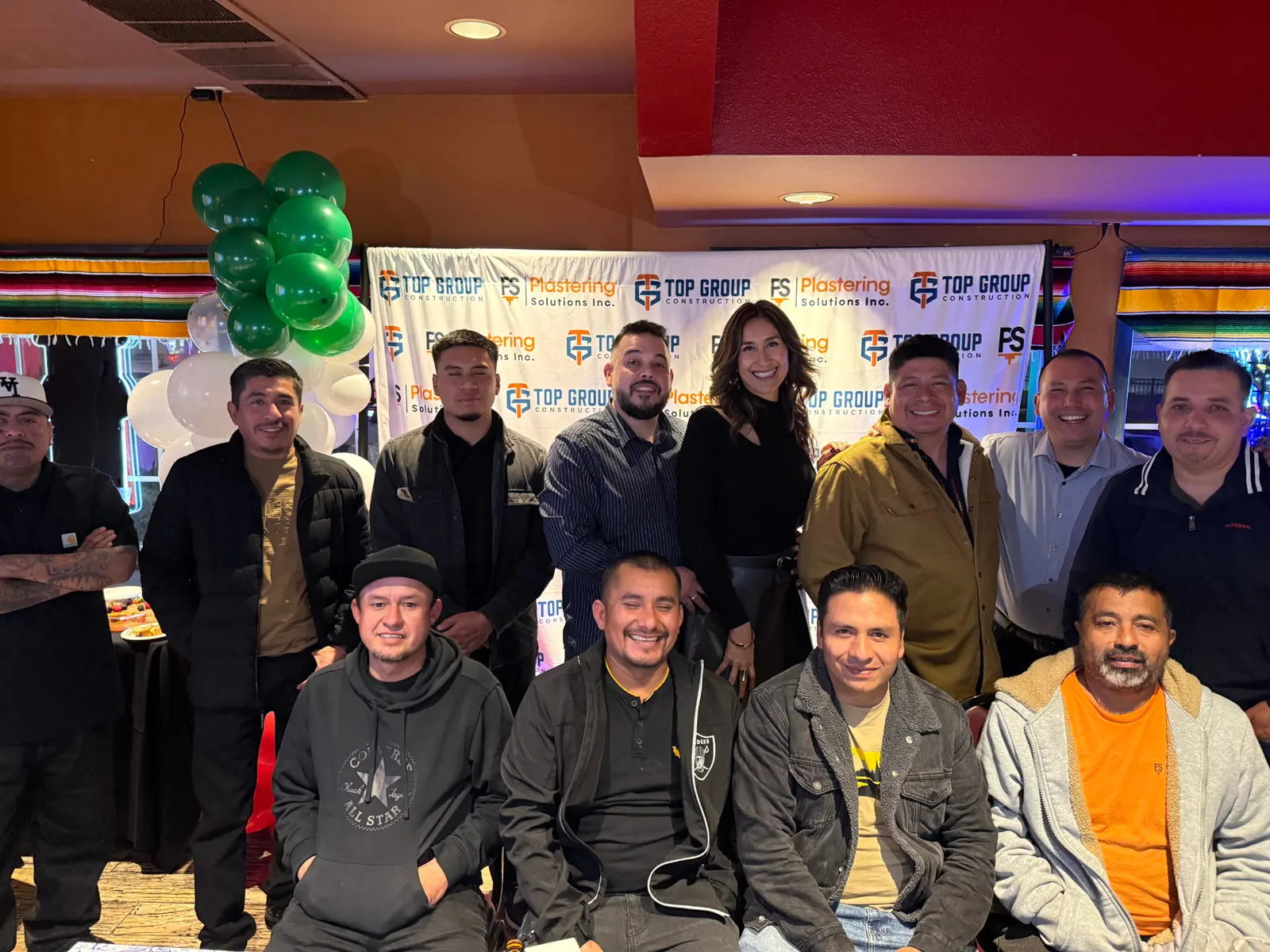 The Plastering Solutions Inc team gathered for an evening event in front of a banner with the Plastering Solutions Inc and Top Group Construction logos, green and white balloons to one side