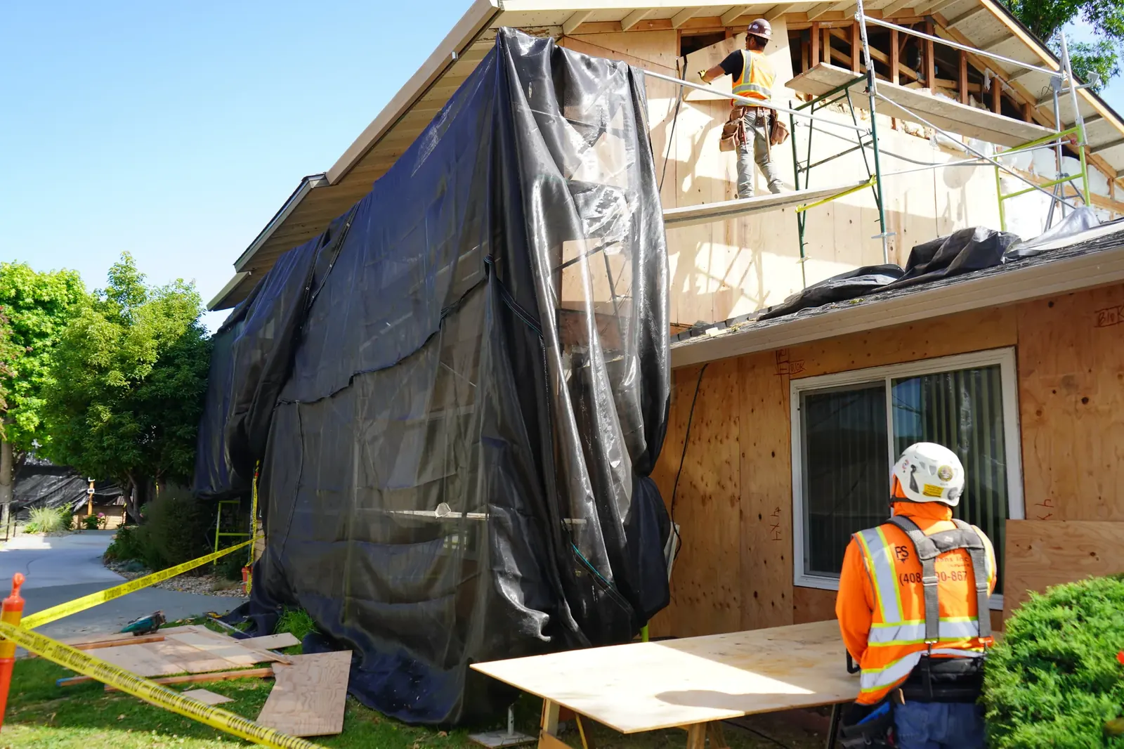 Crew installing plywood sheathing on an upper apartment wall behind scaffold netting